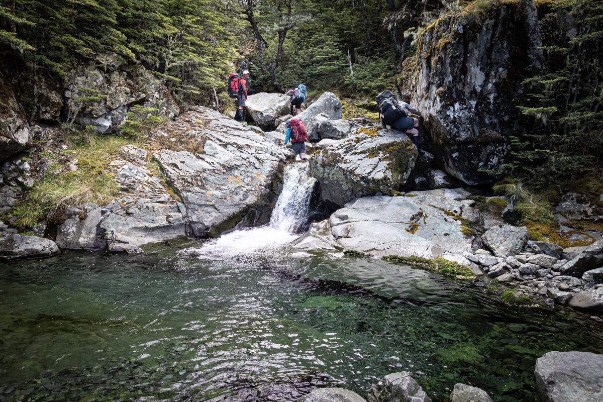 Trampers crossing a small waterfall with a pool below