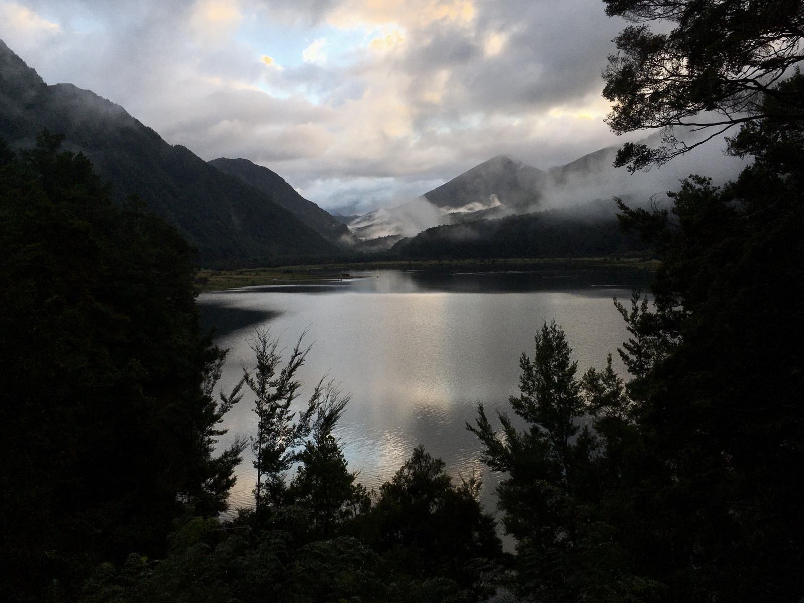 View over a lake with mountains and clouds. Trees in foreground