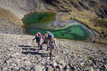 Three trampers ascend scree above a tarn